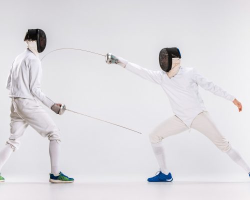 The two men wearing fencing suit practicing with sword against gray studio background
